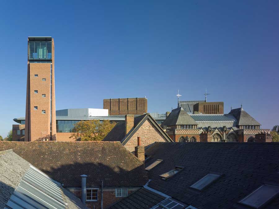 Rooftop view of a mixed architectural complex with a tall brick tower, glass-topped structure, and slate-roofed building in warm daylight tones