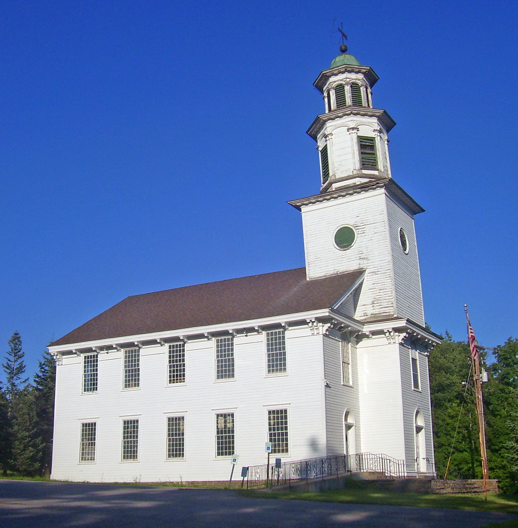 White wooden church building with a tall steeple and green-domed cupola against a clear blue sky